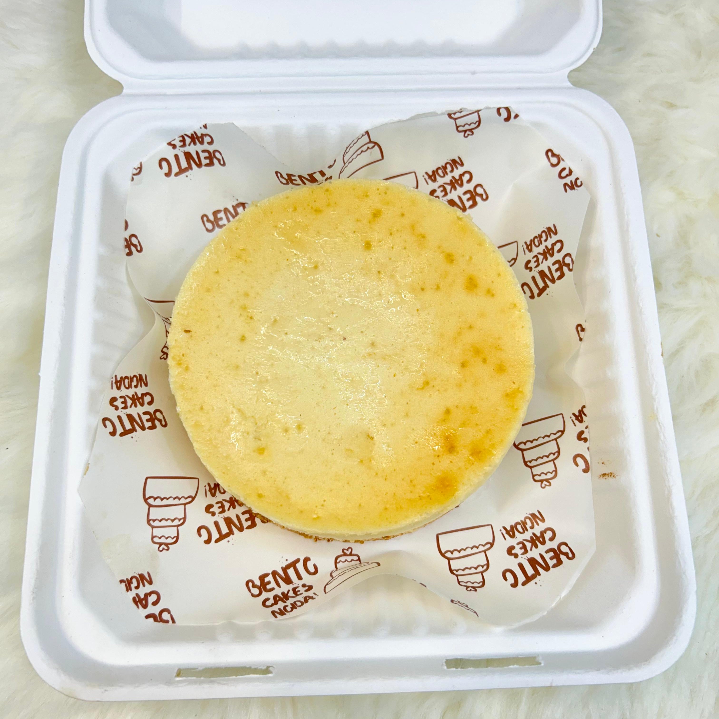 A New York cheesecake in a sleek white bento box, placed on branded parchment paper with ‘BENTO CAKES NOGA’ printed, showcasing its creamy texture and golden crust on a soft white surface with top view.